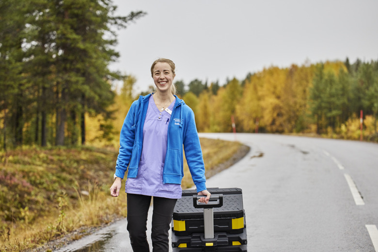 Sjuksköterskan Evelina Markström går på en bilväg med sin stora vårdväska på hjul. Bakgrunden är träd i olika höstfärger.
