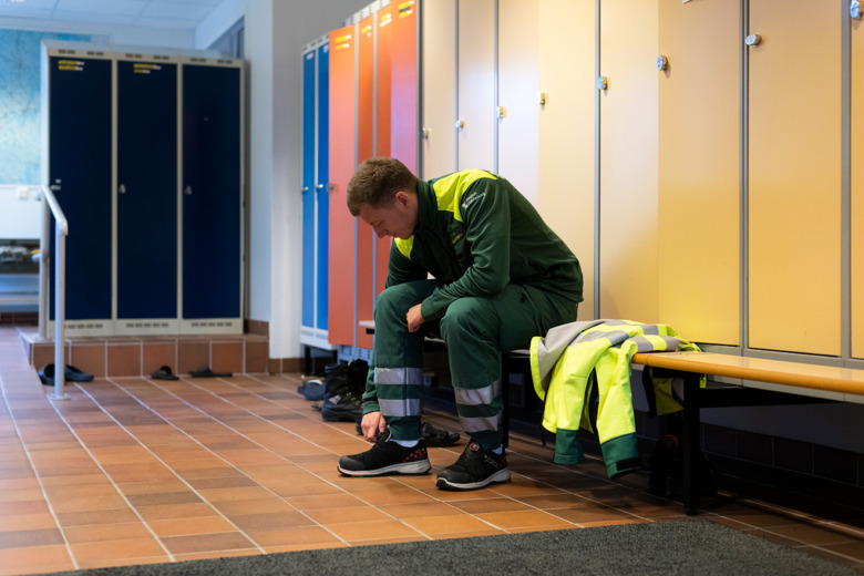 Elias Hedlund sitter i arbetskläder i ett omklädningsrum på sin arbetsplats och sätter på sig sina skor