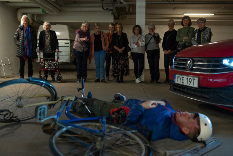 Elva kvinnor och tidigare skolkamrater står på rad och tittar mot en simulering av en cyklist som ligger ner på marken och ska agera skadad efter en krock med en bil. Bredvid cyklisten ligger en cykel och på andra sidan syns fronten av en bil.