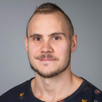 Porträtt av Anton Glans, röntgensjuksköterska. Grå bakgrund, svart mönstrad tröja.