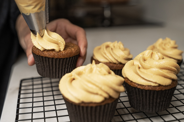 Närbild på fem cup cakes med kolafärgad glasyr av färskost och dulce de leche. Den femte, längst till vänster, håller på att spritsas av Lina Shawali.