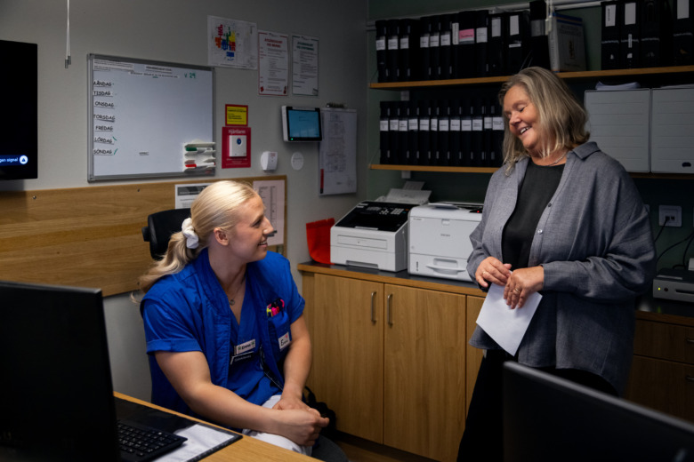 Emma Hall sitter vid ett skrivbord, vänd mot sin chef Ulrika Karlin Gren som står bredvid. De samtalar och ler.