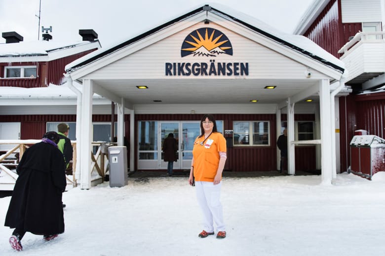 Sjuksköterskan Carina Lantto står utanför entrén till hotell med en stor logga där det står Riksgränsen. Hon har ett stetoskop runt halsen och är iklädd en orange bussarong och vita byxor. Tre personer man bara ser ryggen på är på väg in till hotellet.