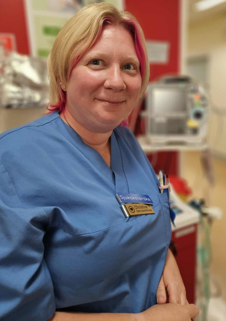 Sjuksköterskan Christine Rommedahl i blå bussarong framför medicinsk-teknisk utrustning i en sjukhuskorridor. Foto: Privat.