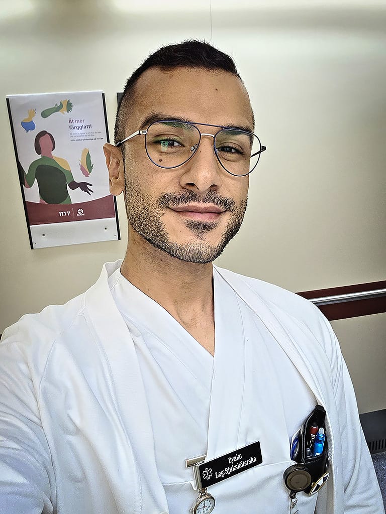 Porträttbild på sjuksköterskan Dyako Ghafoor som är iklädd vita arbetskläder. Bakom honom hänger en skylt från 1177 där det står "Ät mer färgglatt".