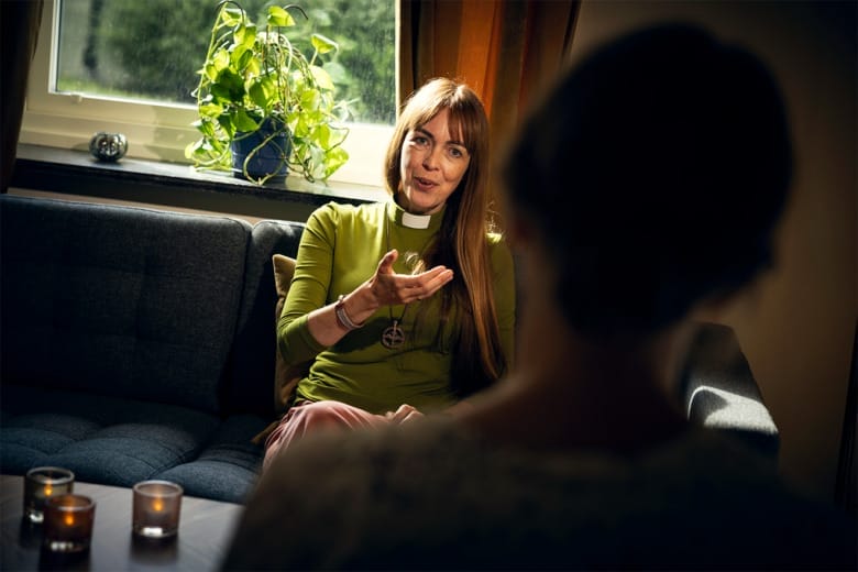 Diakonen Sara Ivarsson sitter i en soffa och samtalar med någon som bara syns som en suddig och mörk figur bakifrån i förgrunden av bilden. Sara håller handen utsträckt mot personen mittemot.