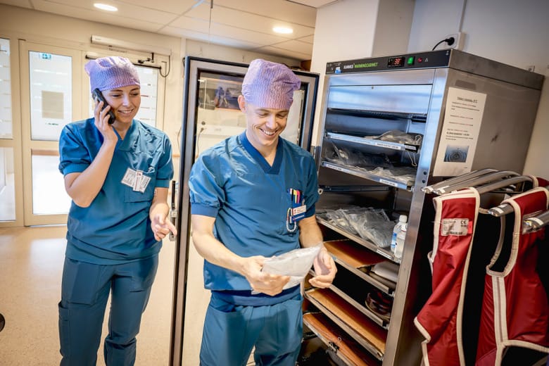 Sjuksköterskan Tobias Ström visar skrattande något han håller i handen för sin kollega som står snett bakom honom. De är båda iklädda blå arbetskläder och varsin operationsmössa.