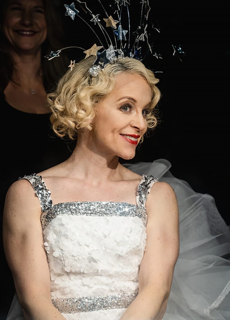 Sjuksköterskan Elin Bemark sminkad med rött läppstift och iklädd scenkläder, en vit klänning med silverdetaljer och vitt tyll bakom. På huvudet har hon ett diadem med stjärnor.