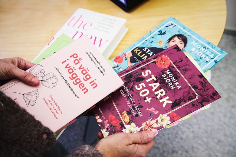 Två händer håller i ett par färgglada böcker. I bakgrunden på ett bord ligger ytterligare böcker. De handlar alla om hormoner, förklimakteriet och liknande ämnen.