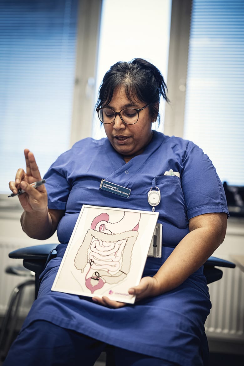 Stomiterapeut Matilda Örn sitter ner och tittar mot en bild som hon håller i. Det är en illustration av magsäcken och tarmarna. Hon gör en gest med handen.