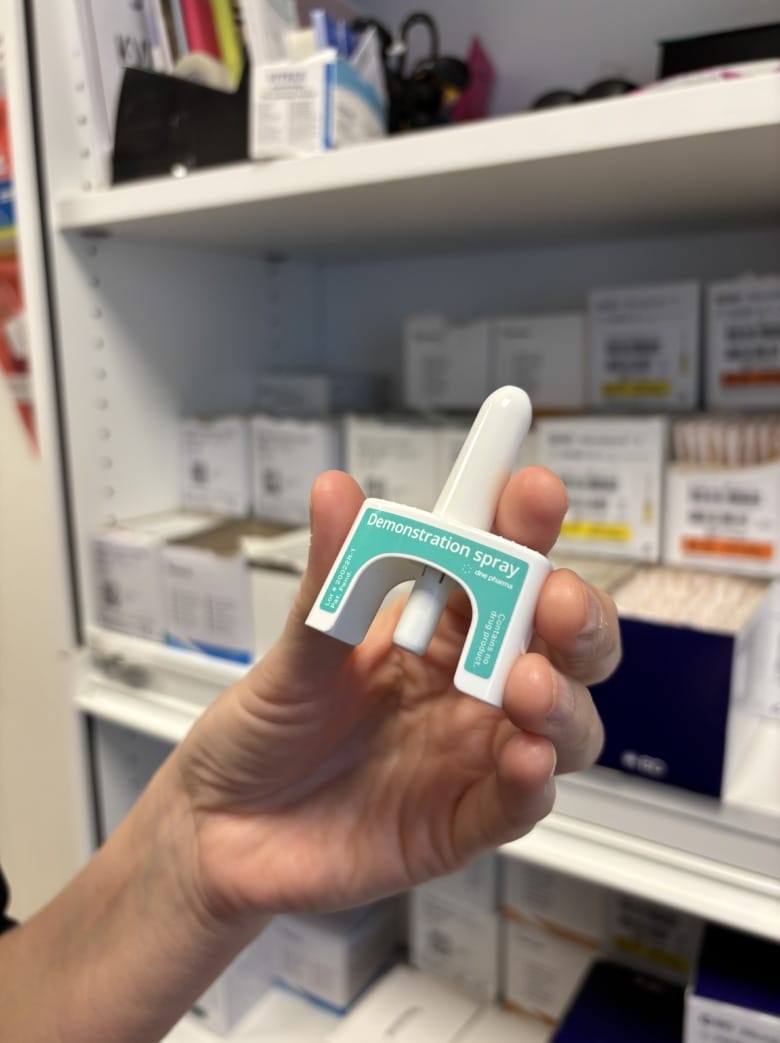 En hand som håller upp en demonstrationsspray med naloxon framför ett skåp med sjukvårdsmaterial i olika lådor.