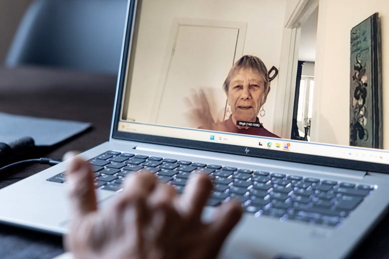 Patienten Birgit Persson syns vinkandes på en datorskärm. I förgrunden syns en hand på tangentbordet som tillhör laptopen.