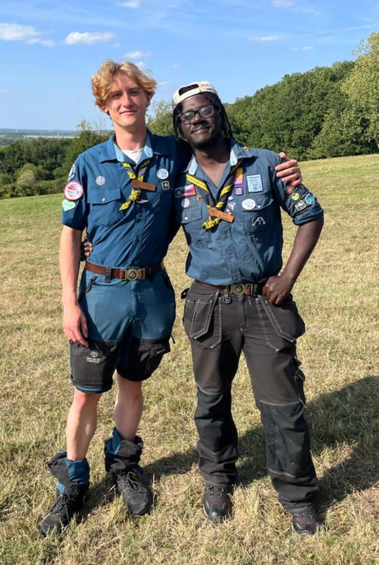 Love Runesson står bredvid en annan ung man och de håller om varandra. De är utomhus på en stor gräsmatta med en klarblå himmel i bakgrunden. De är båda iklädda scoutuniform i form av en klassisk scoutskjorta med tygmärken.
