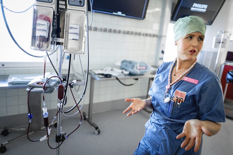 Anestesisjuksköterska Mimmi Ljungholm står iklädd blå arbetskläder och tittar på sidan av kameran och håller ut händerna i en gest. Bredvid henne står en droppställning med blodpåsar kopplade i diverse slangar.