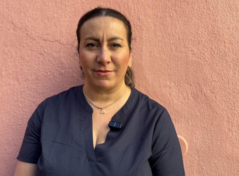 Porträtt på barnmorskan Amneh Elghazzi. Hon har en mörkblå blus och ett guldhalsband med bokstaven A. Står mot ett rosa vägg.