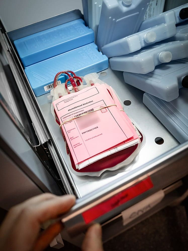 En blodpåse med följesedel i en kyl på operationsavdelningen. En hand drar ut den rostfria hyllan där påsen ligger, bredvid flera blå och grå kylklampar.