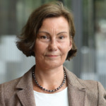 Porträtt av Karin Samuelsson. Hon bär vit topp, beige kavaj, ett halsband med mörtka pärlor och har mörkbrunt hår i pagefrisyr.