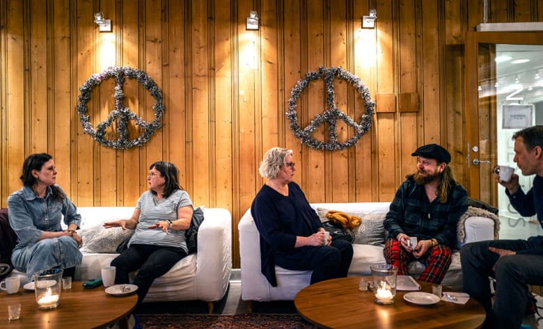 Fyra personer sitter och samtalar i soffor i ett rum med varm belysning. På väggarnas träpaneler hänger peacemärken i form av kransar.