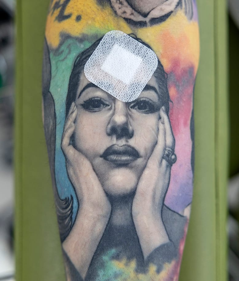 En inzoomad bild på en arm full av tatueringar som bl.a. föreställer operasångerskan Maria Callas. I hennes panna sitter ett plåster efter ett nålstick.