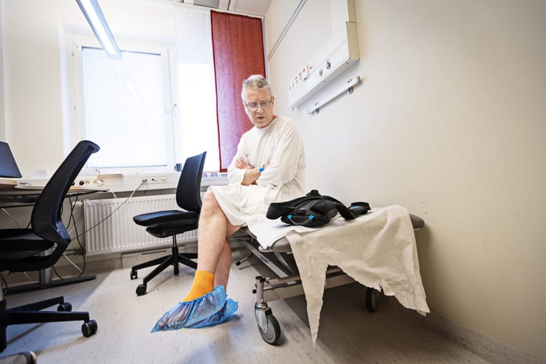Patienten Bengt Danielsson sitter på en brist iförd sjukhuskläder. Han tittar på en svart väska som ligger bredvid honom som håller batterierna till hans hjärtpump.