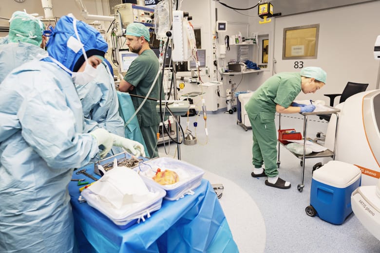En operationssal där flera personer står vid operationsbordet medan några andra står vid sidan av och gör andra saker. En av dessa antecknar något på ett papper, en annan står med ett organ som ligger i en skål.