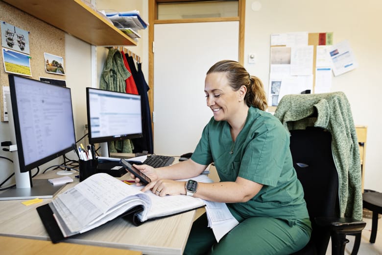 Transplantationskoordinatorn Sofia Ekholm sitter vid ett skrivbord och en dator. Hon iklädd gröna arbetskläder och tittar ner i en pärm på skrivbordet medan hon håller en telefon med ena handen.