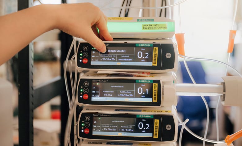 Närbild av tre infusionspumpar staplade på varandra. En hand trycker på den översta pumpens skärm.