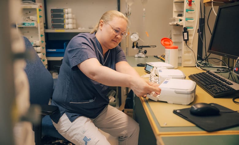 En ingenjör sitter vid ett skrivbord och mekar med en infusionspump. Hon är klädd i sjukhuskläder och jobbar på medicinsktekniska avdelningen på Karolinska sjukhuset. Fler pumpar, en dator och andra instrument syns i bild.