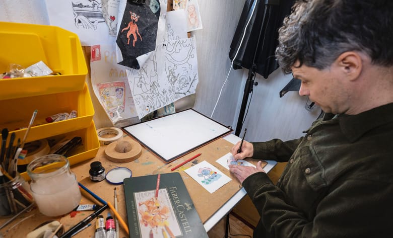 David Henson tecknar i sin ateljé. På bordet ligger färgpennor och tuber och på en anslagstavla sitter flera illustrationer uppsatta.