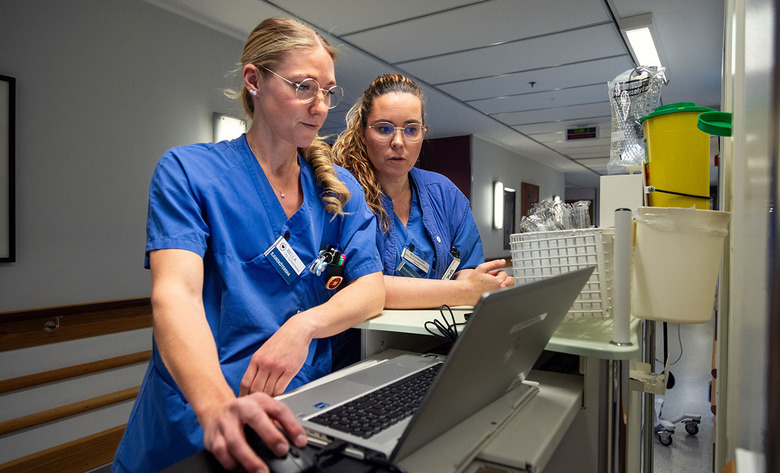 Sjuksköterskorna Isabelle Carles och Alexandra Färnestav står vid en dator och tittar på skärmen. Isabelle har ljust hår i tofs, Alexandra har ljusbrunt uppsatt hår. Båda bär glasögon och blå sjukhuskläder.