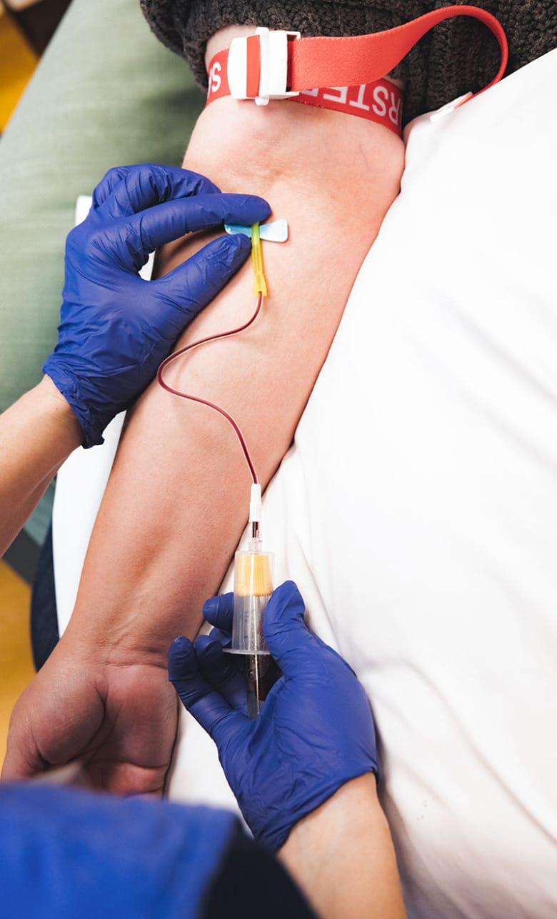 En patient vilar sin arm på en kudde för provtagning. Närbild på sjuksköterskans handskbeklädda händer som sticker patienten i armen.
