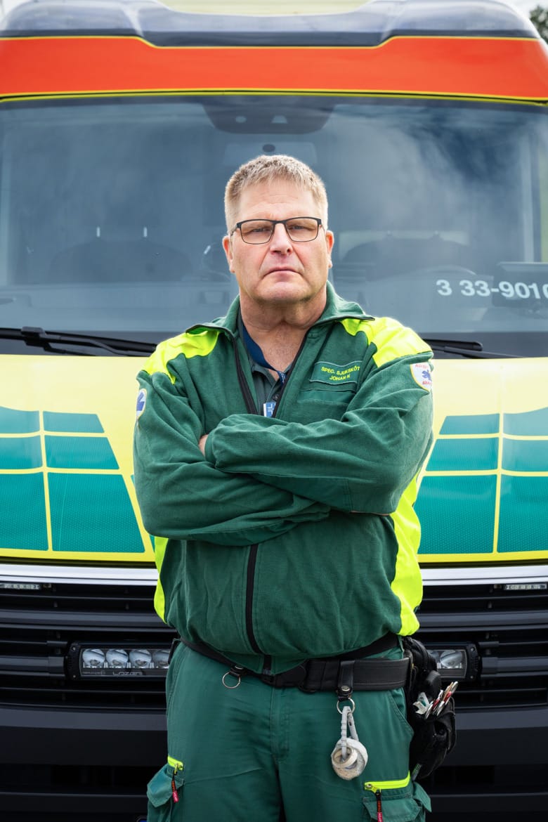En man i gröna ambulanskläder står med armarna i kors framför en ambulans. Porträttet är i halvfigur, mannen bär glasögon och ser bestämd ut.